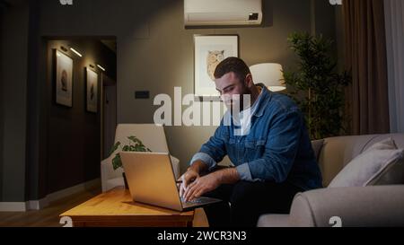 Bärtiger junger Freelancer, der von zu Hause aus mit Laptop arbeitet, während er zu Hause auf dem Sofa sitzt. Ein junger Mann, der auf Bildschirmtippungen am Computer schaut und Arbeit bekommt Stockfoto