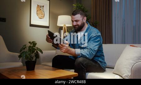 Junger erwachsener bärtiger Mann, der Videospiel auf einem Tablet spielt, während er nachts im Wohnzimmer auf dem Sofa sitzt. Aufgeregt, konzentrierter Mann, der in mich vertieft ist Stockfoto
