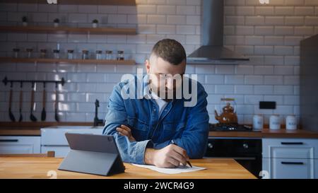 Bärtiger junger erwachsener Mann, der an der Küchentheke sitzt und an einem Tablet arbeitet und Notizen auf Papier macht. Friedliches und entspannendes Arbeiten Stockfoto