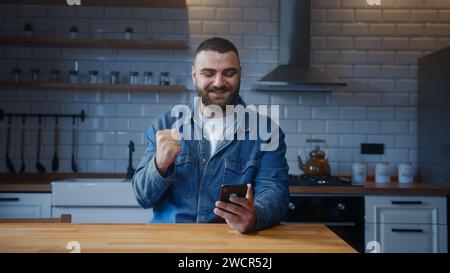 Bärtiger junger Mann, der an der Küchentheke sitzt und Nachrichten liest, erhält gute Nachrichten auf dem Smartphone in E-Mail feiert große Opportun Stockfoto