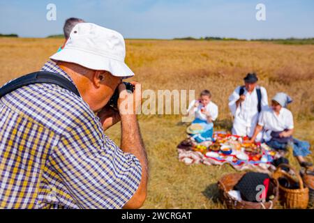 Der Fotograf hält eine Fotokamera, er macht Fotos, betrachtet die Kamera der Familie in weißen Volkstrachten, wie sie im Weizenfeld steht und posiert Stockfoto