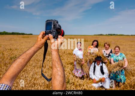 Der Fotograf hält eine Fotokamera, er macht Fotos, betrachtet die Kamera der Familie in weißen Volkstrachten, wie sie im Weizenfeld steht und posiert Stockfoto