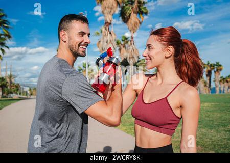 Sportpaare beim Toasten und Trinken von Wasser, während sie sich nach dem Laufen oder Workout ausruhen. Trainer Mann und starke Frau, lächelnd und klirrend ergänzen Flaschen, um eine Pause zu haben und sich während des Trainings zu erfrischen. Hochwertige Fotos Stockfoto