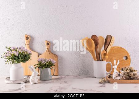 Küchenutensilien aus Holz aus umweltfreundlichen Materialien, Frühlingsblumen in Vase und Kanne, Osterhasenfiguren auf einer Marmorarbeitsfläche. Vorderansicht Stockfoto