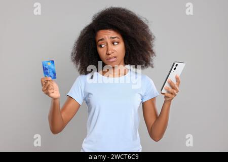 Verwirrte Frau mit Kreditkarte und Smartphone auf grauem Hintergrund. Schuldenproblem Stockfoto