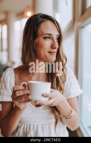 Eine attraktive Frau, die einen Moment am Fenster genießt und eine Tasse frisch gebrühten Kaffee genießt Stockfoto