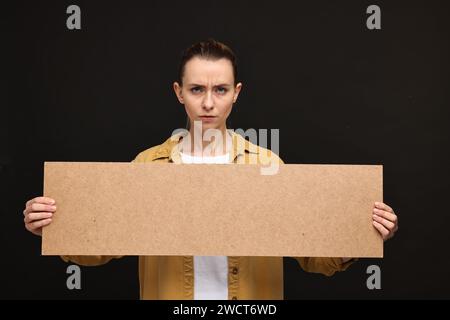 Wütende Frau, die ein leeres Pappbanner auf schwarzem Hintergrund hält, Platz für Text Stockfoto