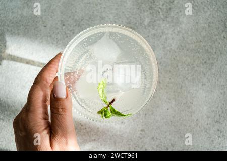 Nahaufnahme eines durchsichtigen Getränks mit Eis, Limettenscheibe und Minze in einem Glasbecher, gehalten von der Hand einer Person über einer grauen Oberfläche Stockfoto