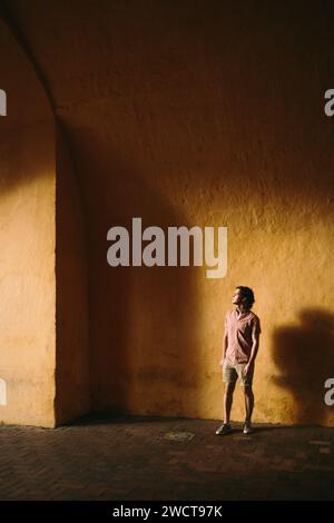 Ein besinnlicher junger Mann steht allein vor einer warmen Wand, die in Sonnenlicht getaucht ist und tiefe Schatten einen starken Kontrast um ihn herum bilden Stockfoto