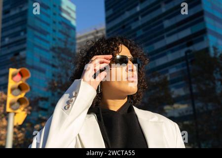 Eine junge, stilvolle Geschäftsfrau passt ihre Sonnenbrille an, während sie im Freien mit Stadtgebäuden und einer Ampel im Hintergrund steht Stockfoto