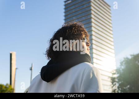 Eine junge Frau blickt nach oben auf ein hohes Gebäude, in sanftem Sonnenlicht getaucht, mit einem nachdenklichen Ausdruck Stockfoto