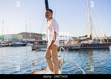 Lächelnder bärtiger Mann in weißem Hemd, der die Yacht vor blauem Meer und Himmel blickt Stockfoto