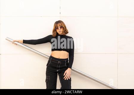 Eine stilvolle junge Frau in einem schwarzen Crop-Top und einer Hose posiert selbstbewusst durch ein silbernes Geländer mit neutralem Wandhintergrund Stockfoto