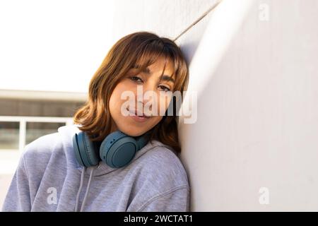 Eine junge Frau lehnt sich an eine Wand, trägt Kopfhörer um den Hals und sonnt sich im Sonnenlicht, mit einem sanften Lächeln im Gesicht Stockfoto