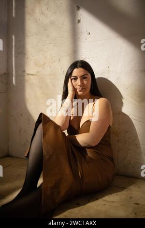 Eine junge Frau in einem schicken Kleid sitzt an einer Wand, das Sonnenlicht wirft dramatische Schatten und ruft eine nachdenkliche Stimmung hervor Stockfoto