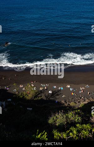 Ein Foto von einem ruhigen schwarzen Sandstrand mit Sonnenbaden und dem ruhigen Meer, das den Kontrast zwischen dem dunklen sa unterstreicht Stockfoto