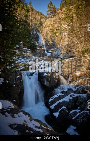 Ein heiterer Bergwasserfall fällt über Felsen, umgeben von einem Wald, der leicht mit Schnee bedeckt ist und sich in sanftem Sonnenlicht sonnt Stockfoto