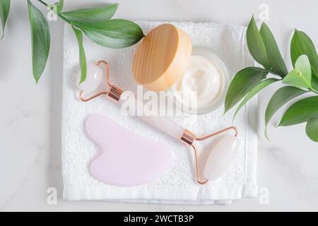 Pflegeprodukte wie Jadewalze, Gua Sha und Feuchtigkeitscreme, die neben grünen Blättern auf einem weißen Handtuch liegen Stockfoto