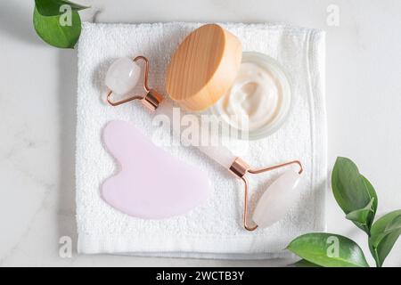 Flache Hautpflegeprodukte mit Jaderolle, Gua Sha und Feuchtigkeitscreme auf einem weichen Handtuch mit grünem Laub Stockfoto