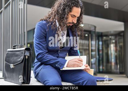 Lateinischer Geschäftsmann mit langen Haaren in einem Notizbuch, der draußen in der Nähe einer Geschäftsaktentasche sitzt Stockfoto