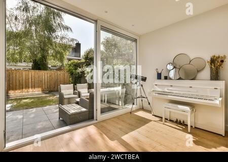 Geräumiges, modernes Wohnzimmer mit Tageslicht, einem weißen Klavier und einer offenen Schiebetür, die in einen Garten mit Sitzgelegenheiten im Freien führt. Stockfoto