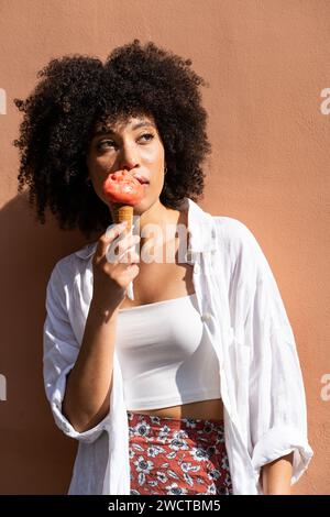 Schwarze Frau mit lockigen Haaren in weiß genießt eine Erdbeere-Eiscreme vor einer warmen Pfirsichwand und blickt weg Stockfoto