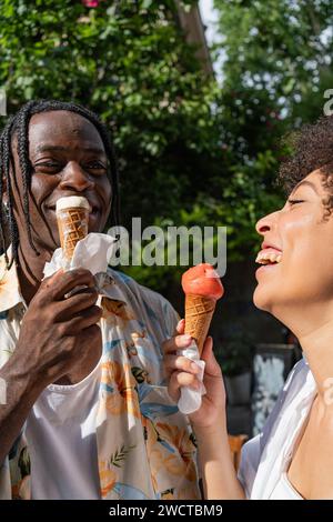 Ein fröhliches Paar schwarzer Freunde, die an einem sonnigen Tag mit grünem Laub im Hintergrund mit Eiskegel lachen Stockfoto