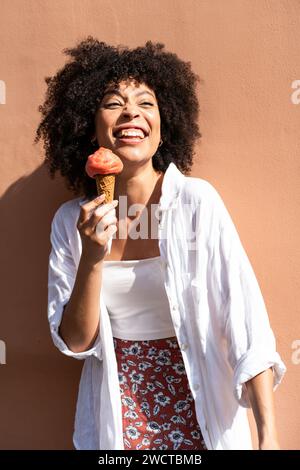 Schwarze Frau mit lockigen Haaren in weiß genießt eine Erdbeere-Eiscreme vor einer warmen Pfirsichwand und blickt weg Stockfoto