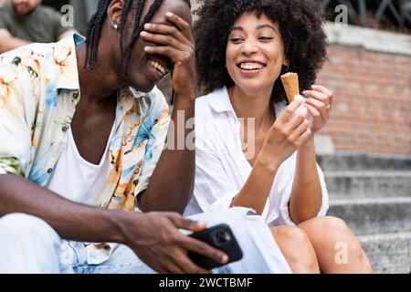 Freunde lachen auf Steintreppen, während einer eine Eiskegel hält und einen offenen Moment festhält Stockfoto