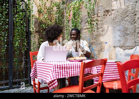 Ein schwarzer Mann und eine schwarze Frau genießen eine Mahlzeit zusammen mit einem Laptop auf dem Tisch in einer bezaubernden Umgebung im Freien Stockfoto
