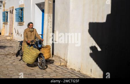 Ein bärtiger tunesischer Mann mit Ohrstöpseln trägt große handgewebte Taschen auf seinem Motorrad durch eine Kopfsteinpflasterstraße in der alten Medina von Sousse in Tunis Stockfoto