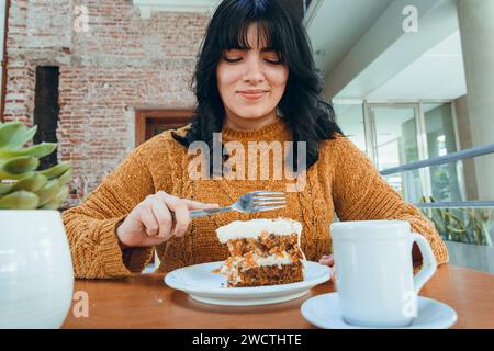 Porträt einer jungen lateinfrau mit schwarzen Haaren und braunem Pullover, drinnen sehr glücklich sitzend, Karottenkuchen mit Kaffee im Restaurant, Gebäck Stockfoto