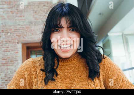 Porträt einer jungen lateinamerikanischen venezolanischen Frau mit schwarzen Haaren und braunem Pullover, überrascht, wie sie mit einem großen glücklichen Lächeln in die Kamera blickt. Stockfoto