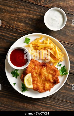 Hausgemachtes paniertes Schnitzel mit Kartoffelfritten und Sauce auf weißem Teller auf hölzernem Hintergrund. Draufsicht, flach Stockfoto