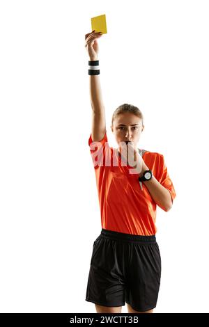 Junge ernste Frau, Fußballschiedsrichter, der eine Pfeife bläst und gelbe Karte als Warnsymbol vor weißem Studiohintergrund zeigt Stockfoto