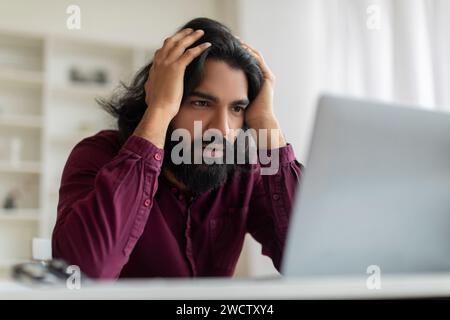 Besorgter indischer Mann, der frustriert den Kopf fasst, während er auf den Computerbildschirm starrt Stockfoto