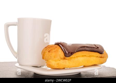 Ein süßer Schokoladeneklair auf einer weißen Keramikuntertasse mit einer Tasse auf einem Schieferstein, Nahaufnahme isoliert auf weißem Hintergrund. Stockfoto