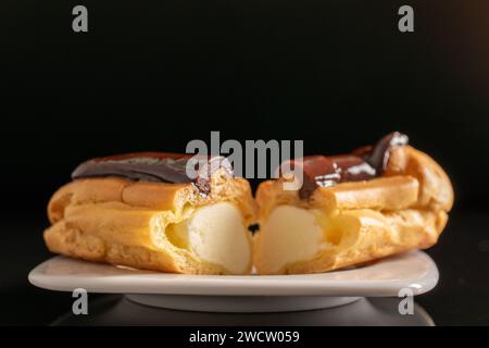 Zwei Hälften einer köstlichen Schokoladeneklair auf einer weißen Keramikplatte, Nahaufnahme isoliert auf schwarzem Hintergrund. Stockfoto