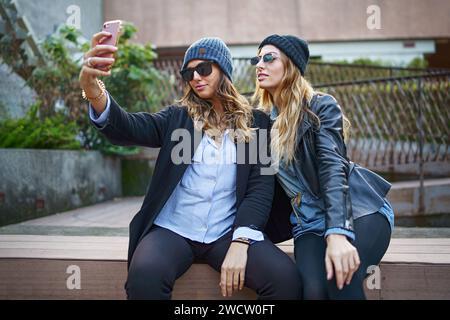 Zwei modische junge Frauen in Freizeitkleidung und Mützen machen ein Selfie mit einem Smartphone, während sie draußen sitzen und Freundschaft und modernes Leben zeigen Stockfoto