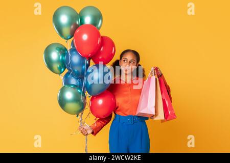 afrikanisches Teenager-Mädchen hält Einkaufstaschen und Ballons vor gelbem Hintergrund Stockfoto