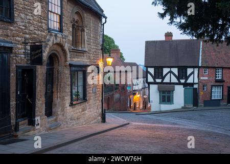 Lincoln, Lincolnshire, England. Blick auf den kopfsteingepflasterten steilen Hügel an der Kreuzung mit Michaelgate, Dämmerung, Straßenlaternen, die historische Gebäude beleuchten. Stockfoto