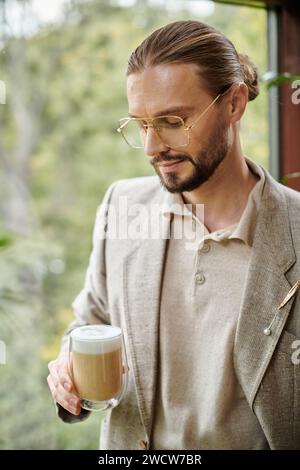 Attraktiver ansprechender Mann mit Bart und gesammelten Haaren in elegantem Anzug, der seinen heißen Kaffee trinkt Stockfoto