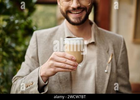 Gut aussehender glücklicher Mann mit Bart und gesammelten Haaren in elegantem Anzug, der seinen heißen Kaffee genießt Stockfoto