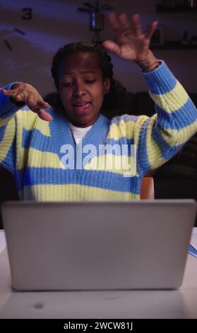 Fröhliche Afroamerikanerin, die nachts vor dem Laptop sitzt und im Büro singt und tanzt. Stockfoto