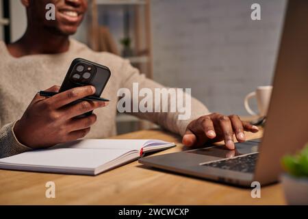 Glücklicher afroamerikanischer Mann mit Smartphone, der bei der Arbeit zu Hause ein Notebook benutzt, zugeschnittene Ansicht Stockfoto