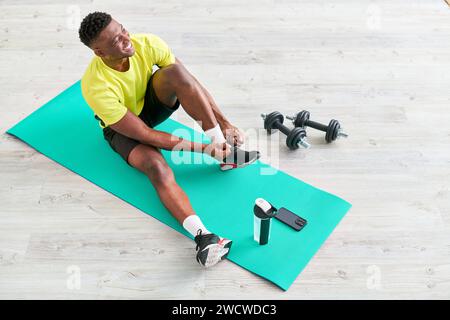 Lächelnder afroamerikanischer Mann, der Sneaker auf einer Fitnessmatte in der Nähe von Smartphone und Sportgeräten schnürt Stockfoto