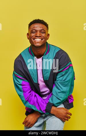Überfröhlicher und stilvoller afroamerikanischer Mann in heller Windjacke mit Blick auf die Kamera auf Gelb Stockfoto
