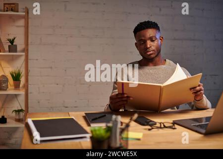 Junger, nachdenklicher afroamerikanischer Freelancer, der sich bei der Arbeit zu Hause nachts ein Notebook anschaut Stockfoto