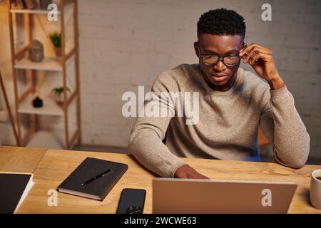 Ernsthafter after afroamerikanischer Freelancer mit Brillen, der in der Nähe eines Laptops denkt und nachts von zu Hause aus arbeitet Stockfoto