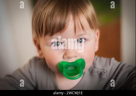 Der süße Junge blickt direkt in die Kamera, die Neugier in seinen Augen, mit einem leuchtend grünen Schnuller im Mund vor einem sanft fokussierten Innenraum Stockfoto